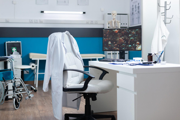 Medical exam room with wheelchair, desk, and equipment setup.